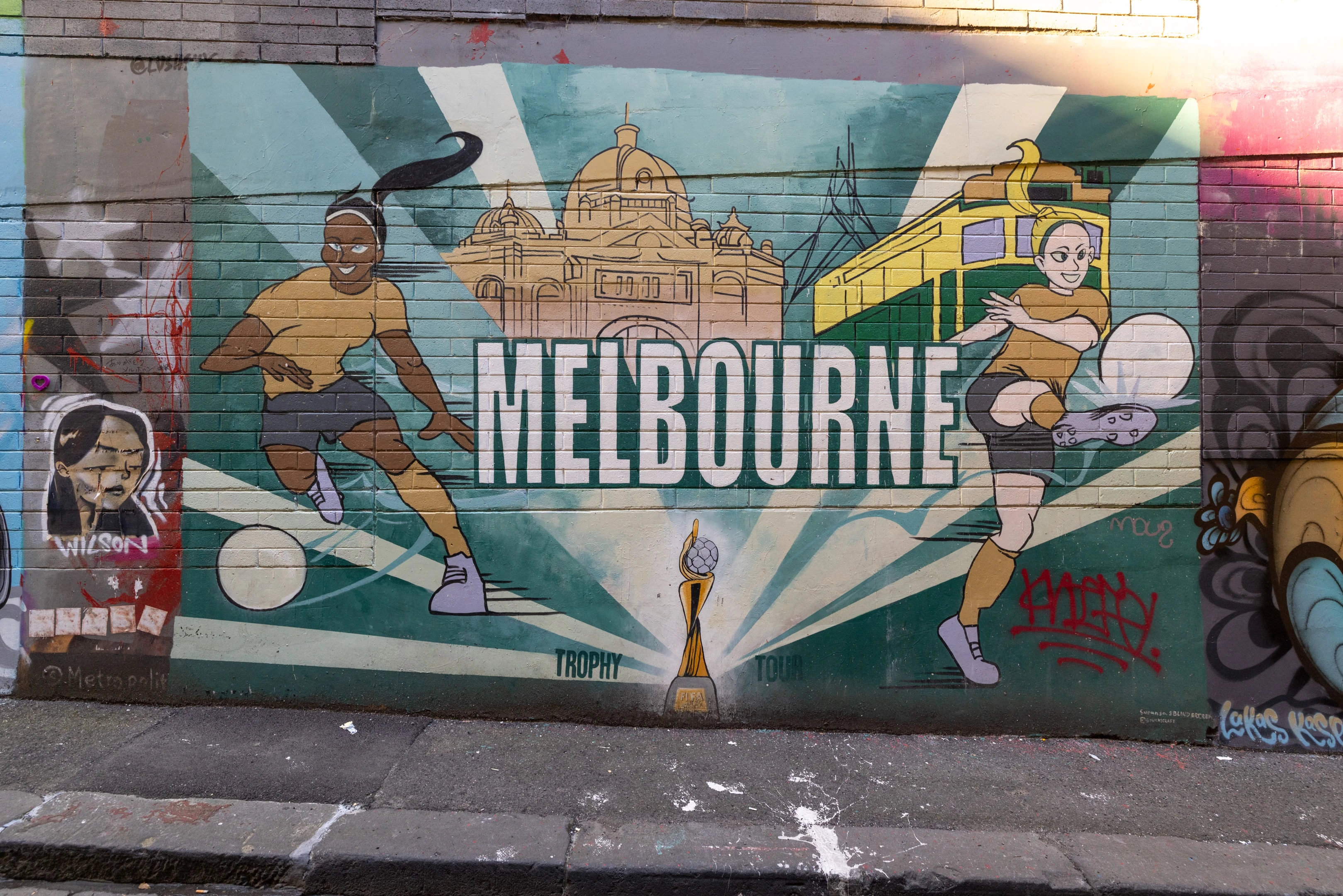 A work of street art containing the word Melbourne in capitals and depicting two women kicking footballs, Flinders Street station and a tram